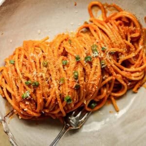 A close-up of a bowl of vegan gochujang vodka pasta coated in a rich red sauce, garnished with grated cheese and chopped green herbs. A fork rests beside the pasta on the white ceramic plate.